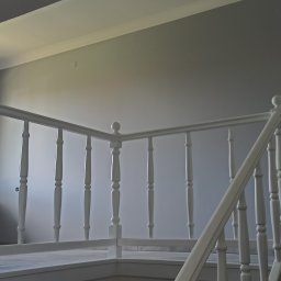 Łukasz Stępień - Klatka schodowa z białymi balustradami w jasnym, nowo pomalowanym wnętrzu domu; widoczny grzejnik pod oknem.