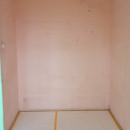 Łukasz Stępień - Pokój przygotowany do malowania: podłoga zabezpieczona folią i taśmą malarską, ściany w odcieniach różu, widoczne ślady po wcześniejszych pracach.