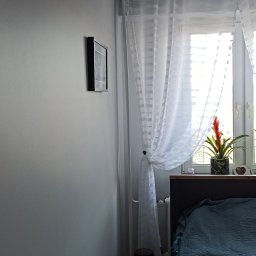 Łukasz Stępień - Szara ściana w pokoju w Rzeszowie po świeżym malowaniu, widoczna listwa sufitowa, obrazek w czarnej ramce, białe firanki w oknie i fragment łóżka z niebieską narzutą.