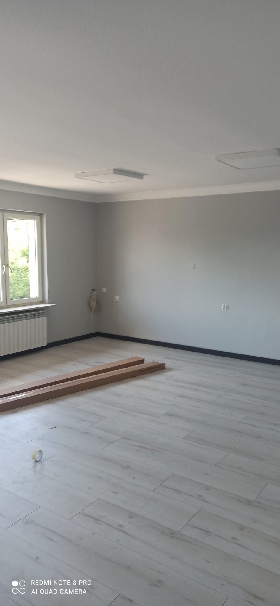 Pomalowane na szaro wnętrze pokoju z ułożonymi panelami podłogowymi i przygotowanymi panelami do montażu, widoczne także grzejnik, okno i dwa kwadratowe panele oświetleniowe na suficie.