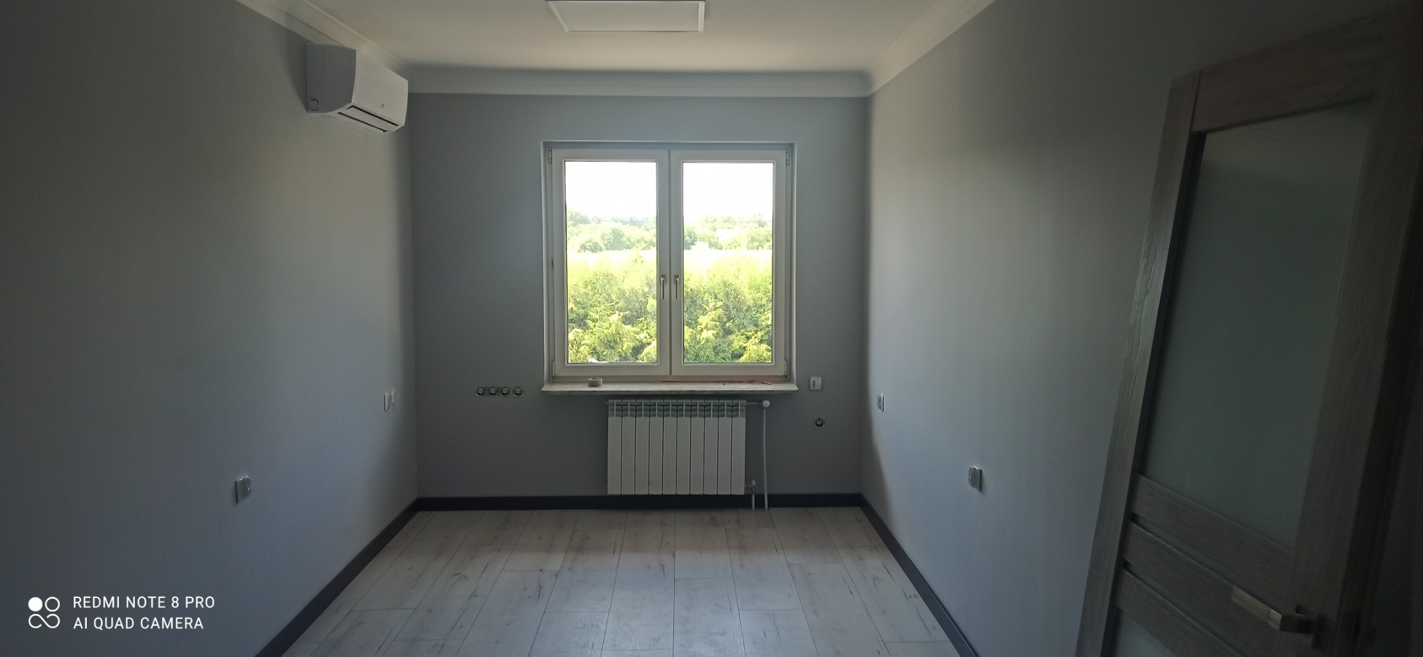 Pomalowane na szaro wnętrze pokoju z panelami podłogowymi, grzejnikiem pod oknem i klimatyzacją na ścianie.