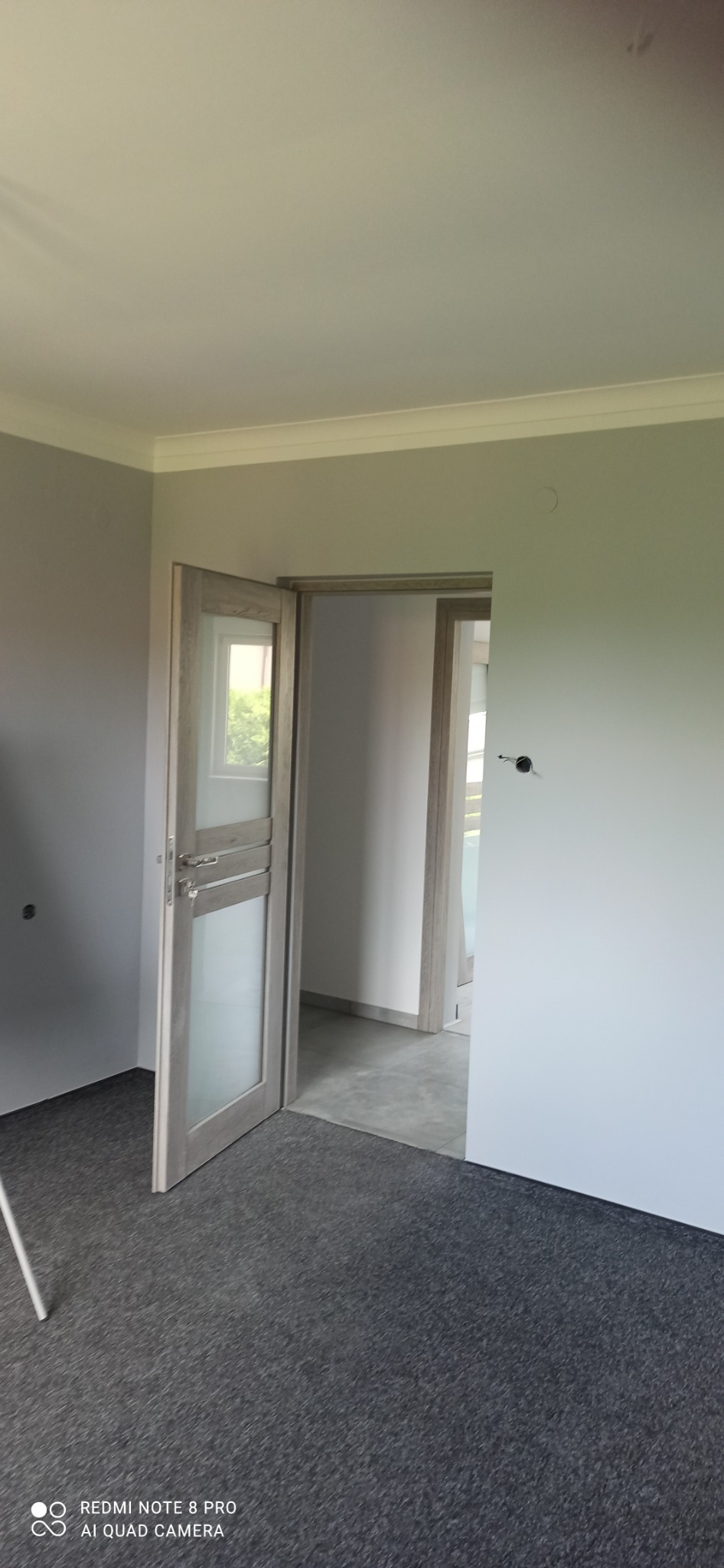 Wykończone wnętrze pokoju z szarą wykładziną i częściowo otwartymi, przeszklonymi drzwiami w odcieniach szarości.