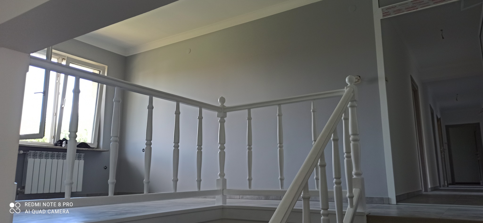 Klatka schodowa z białymi balustradami w jasnym, nowo pomalowanym wnętrzu domu; widoczny grzejnik pod oknem.