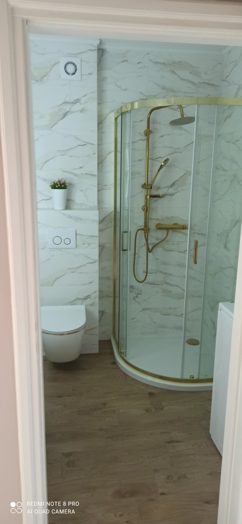 Nowoczesna łazienka z kabiną prysznicową ze złotymi elementami, marmurowe płytki ścienne, podwieszana toaleta i drewnopodobna podłoga, widok zza framugi drzwi.