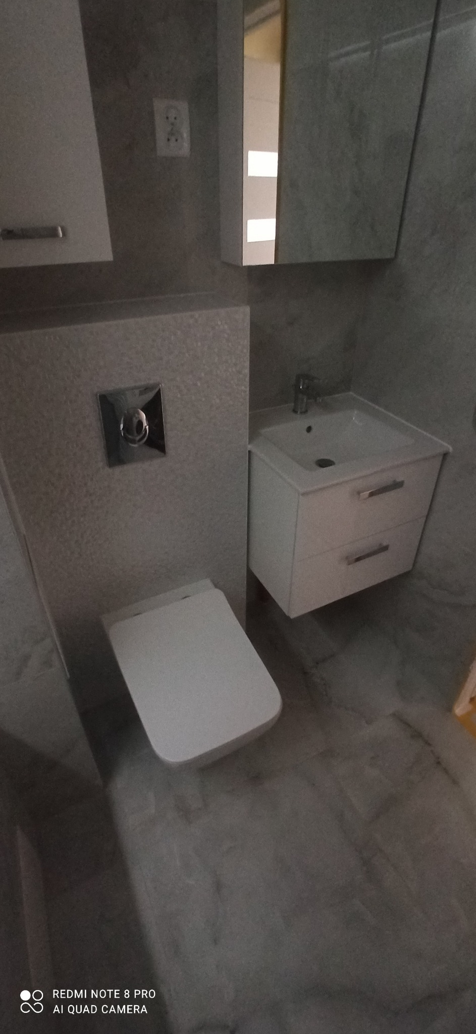 Nowoczesna łazienka z wiszącą toaletą, umywalką z szafką i szarą płytką imitującą kamień.