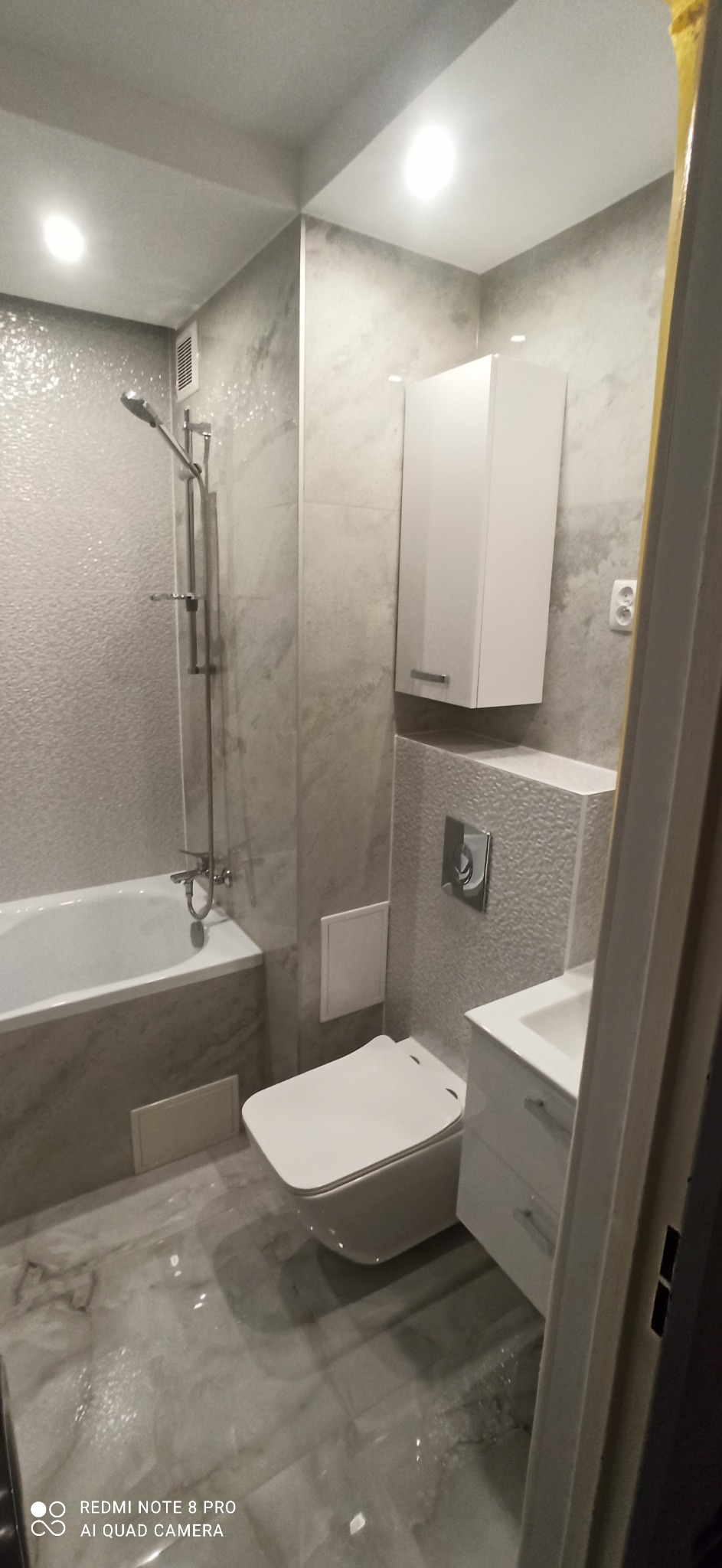 Nowoczesna łazienka z szarymi płytkami imitującymi marmur, wisząca toaleta, biała szafka nad stelażem podtynkowym, biała umywalka z szafką i wanna z chromowaną armaturą.