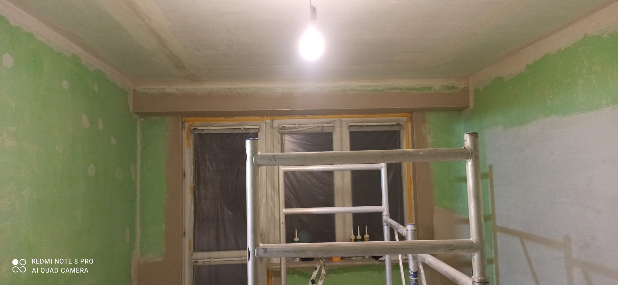 Pokój w trakcie przygotowania do malowania: ściany częściowo pokryte szpachlą, zabezpieczone okna, rusztowanie w pomieszczeniu.