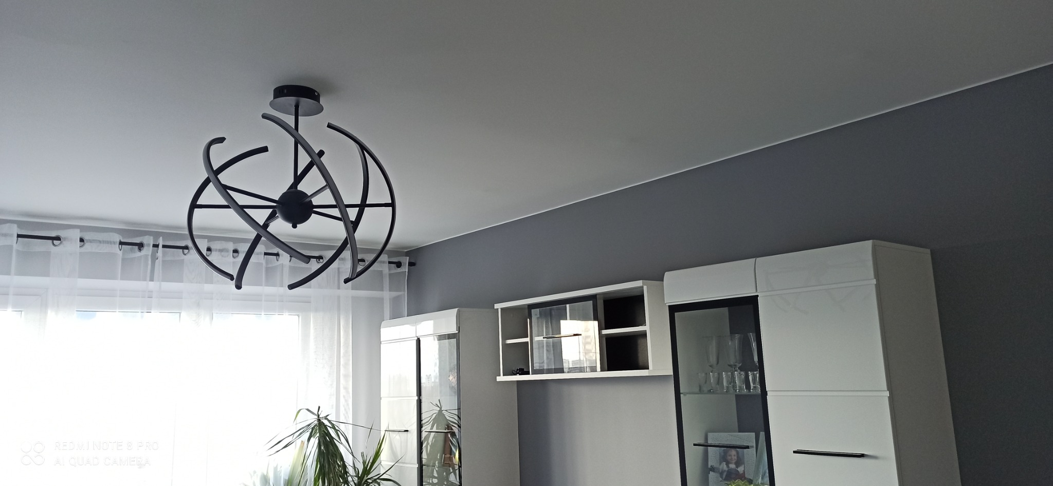 Nowoczesna lampa sufitowa w kształcie orbit na tle szarej ściany i białych mebli w salonie.