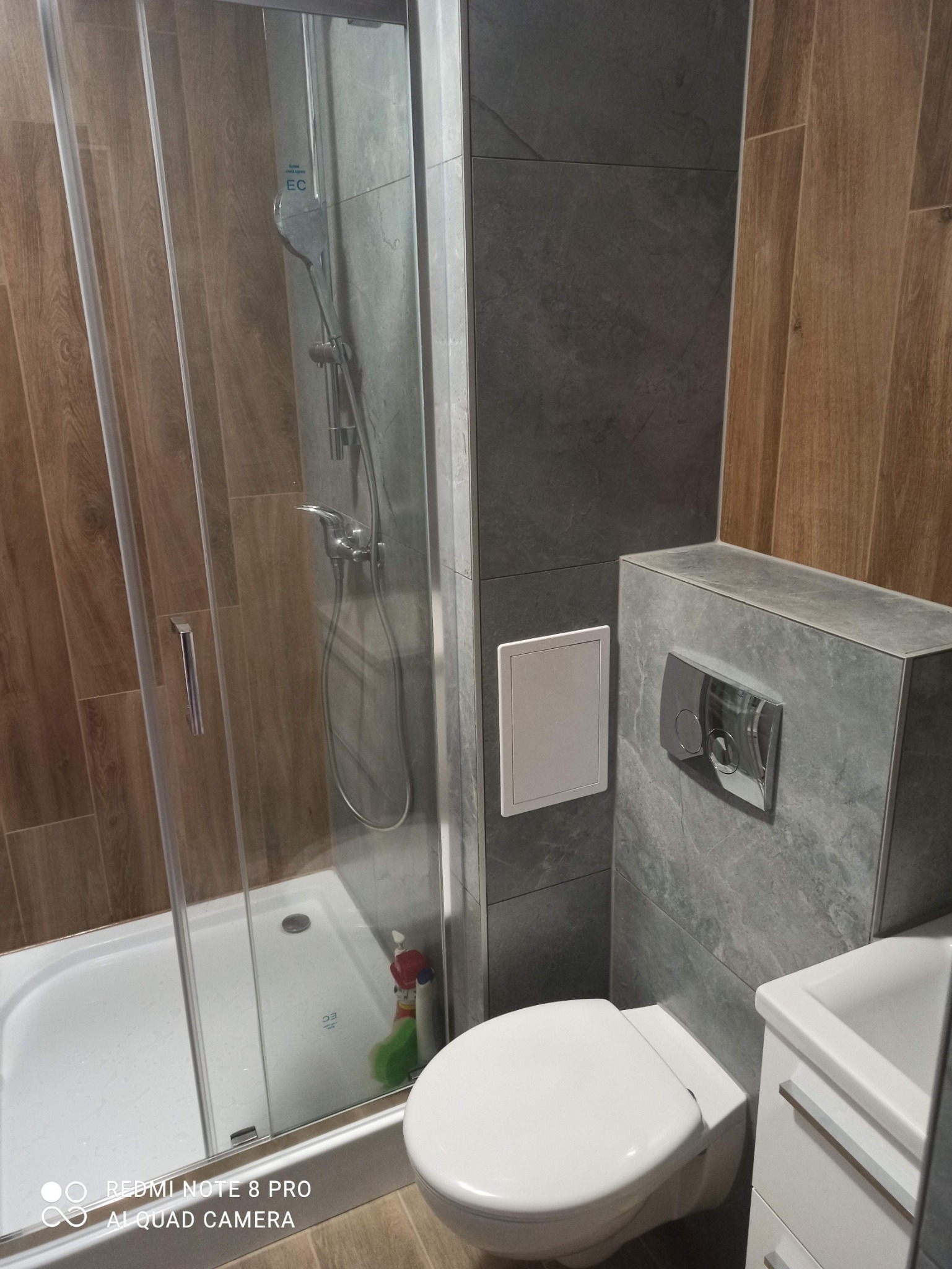 Wykończona łazienka z kabiną prysznicową i wiszącą toaletą, szare płytki imitujące kamień na ścianach, drewnopodobne płytki na fragmencie ściany, biała zabudowa umywalki.