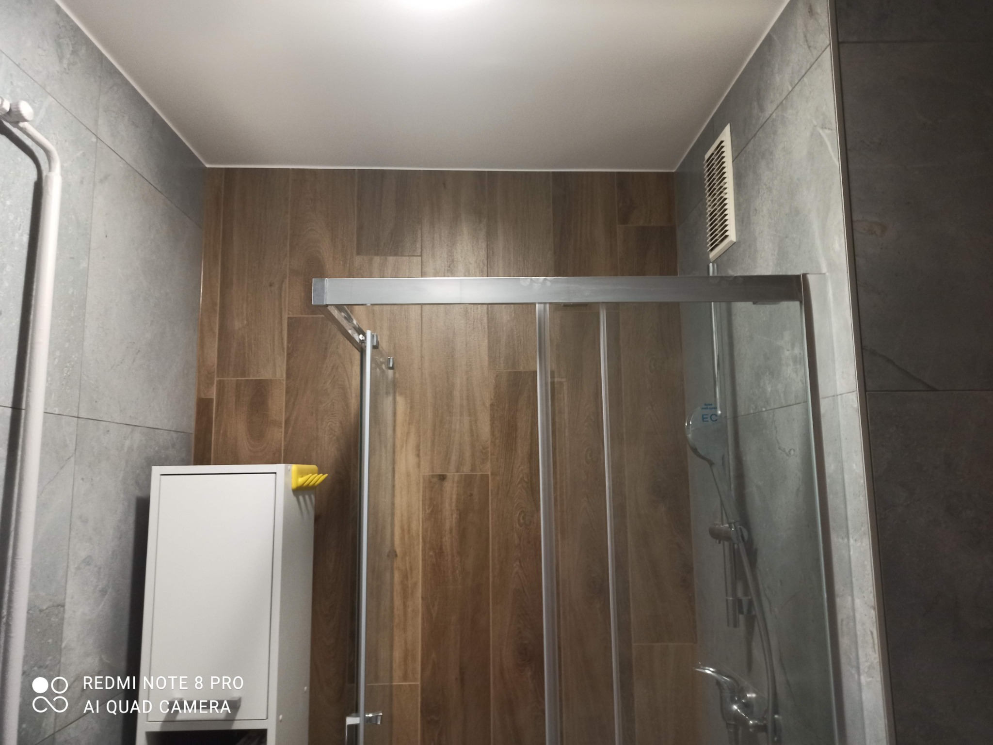 Nowoczesna kabina prysznicowa ze szklanymi drzwiami na tle płytek imitujących drewno i szarych płytek, z białą szafką z żółtym uchwytem na ręczniki po lewej stronie.