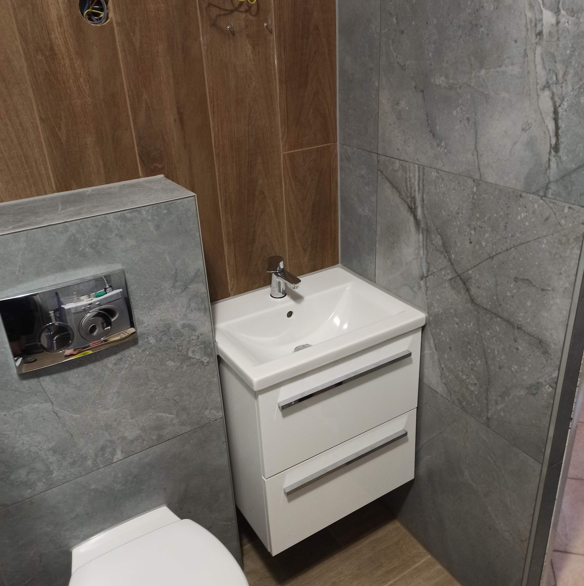 Nowoczesna łazienka po remoncie: szare płytki imitujące kamień, drewnopodobne panele ścienne, biała szafka pod umywalką z chromowaną baterią i podwieszany sedes z chromowanym panelem spłukującym.