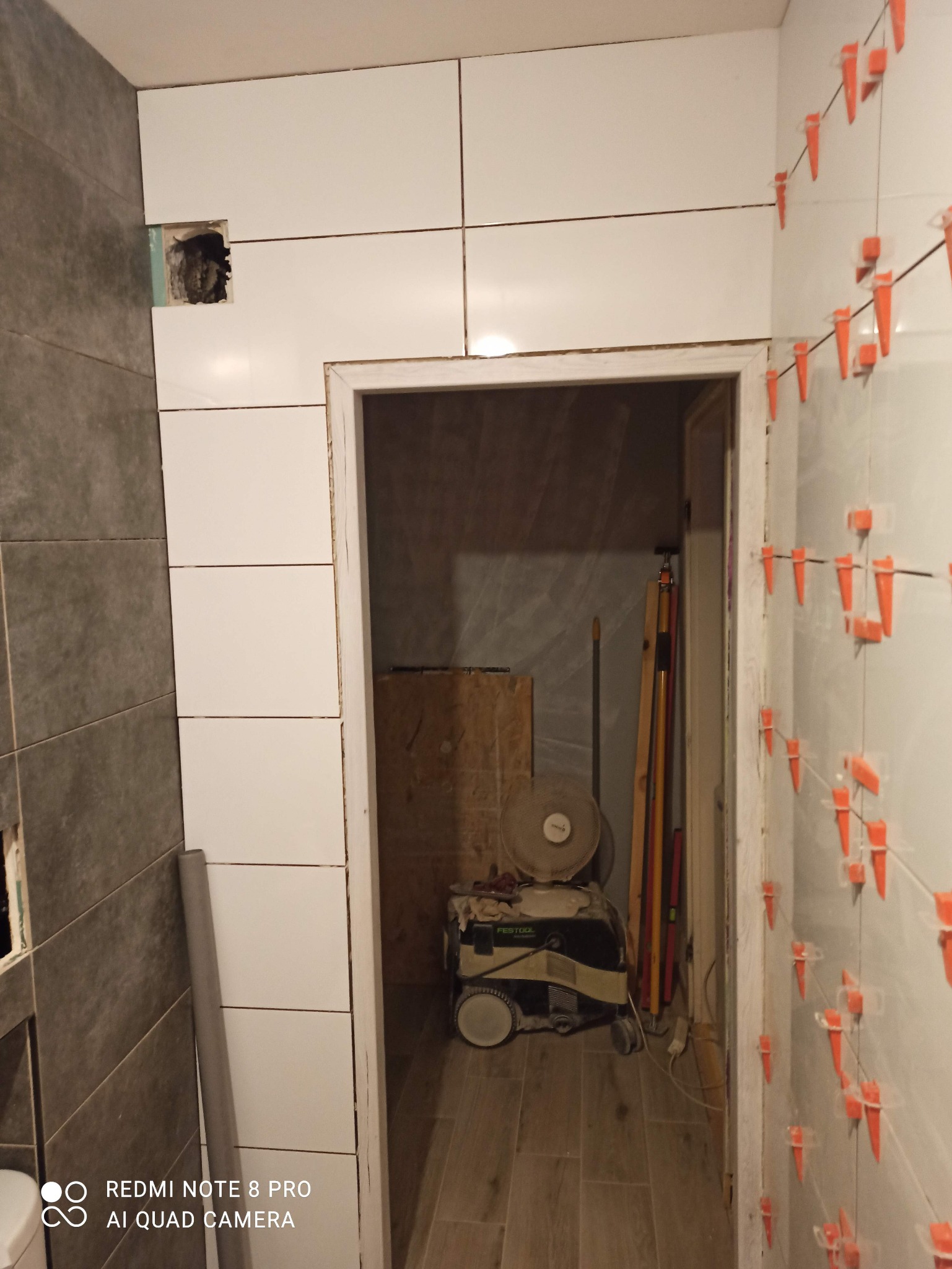 Proces układania białych, prostokątnych płytek na ścianie, widoczne pomarańczowe krzyżyki dystansowe, w tle widoczny sprzęt budowlany i wentylator, otwór w ścianie bez płytek.