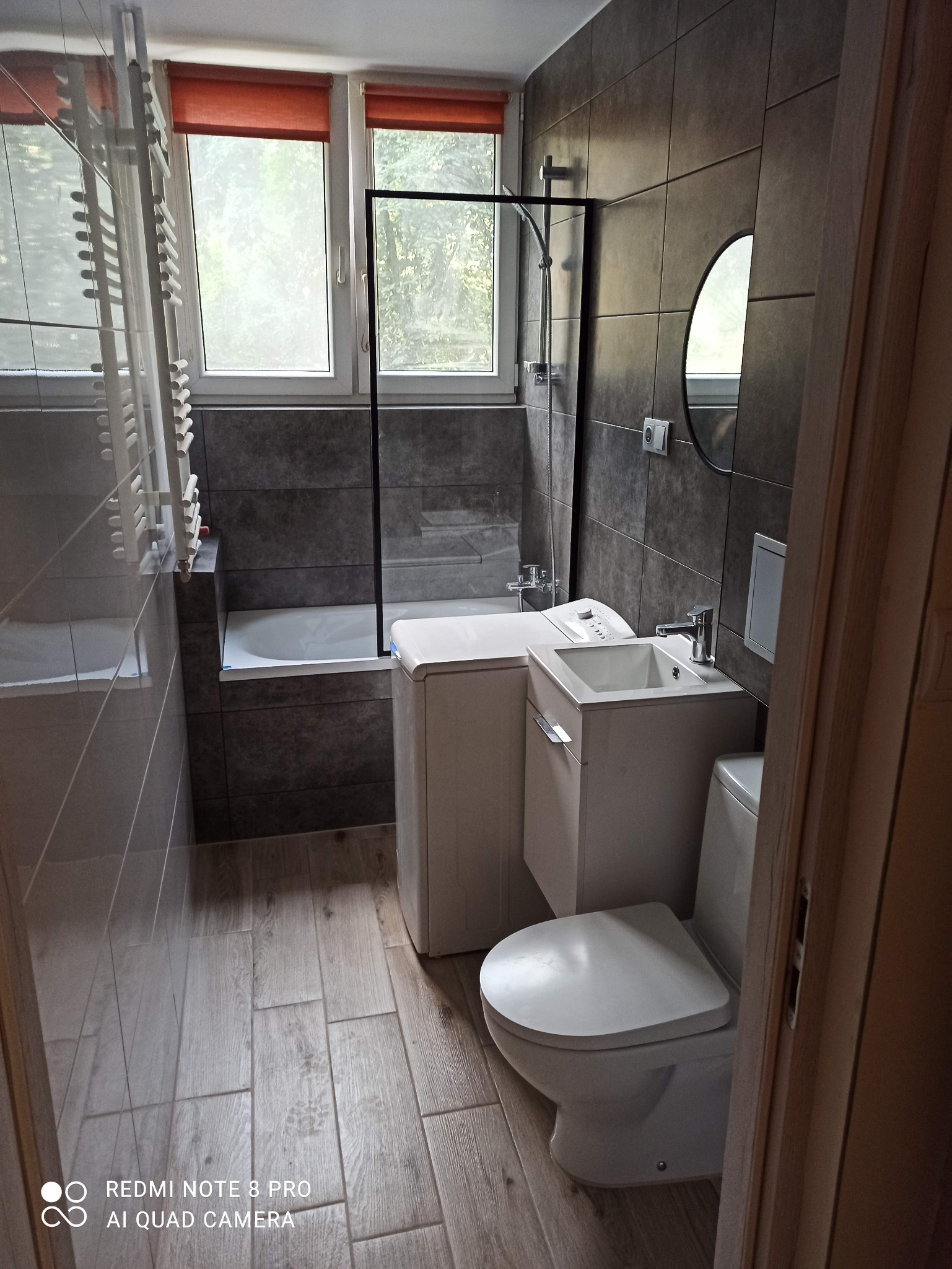 Ukończona łazienka z ciemnymi płytkami, drewnopodobną podłogą, umywalką z szafką, toaletą, wanną za szklaną ścianką i pralką ładowaną od góry.