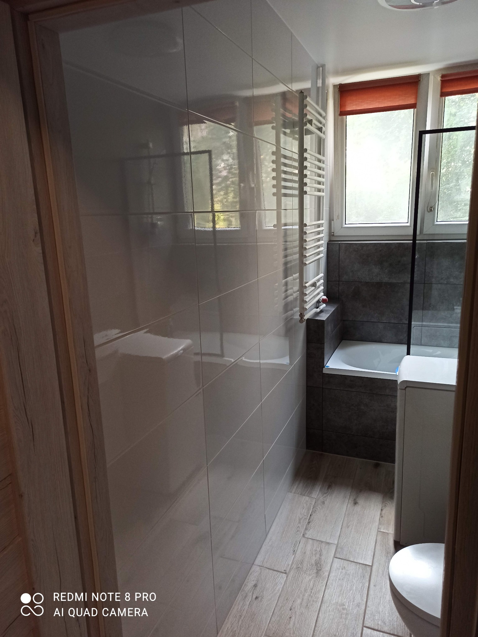 Nowoczesna łazienka po remoncie: białe, błyszczące płytki ścienne, szare płytki imitujące kamień przy wannie, drewnopodobne płytki podłogowe, grzejnik drabinkowy i okno z pomarańczowymi roletami.