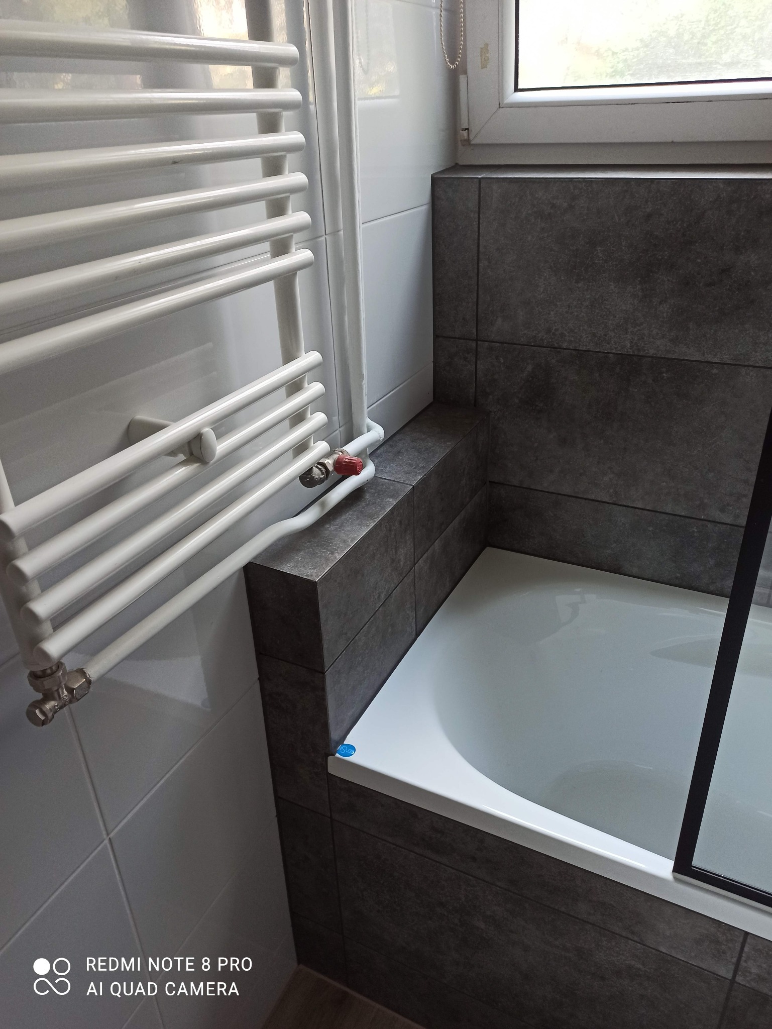 Biała wanna obudowana ciemnoszarą glazurą, grzejnik drabinkowy na ścianie z białych płytek, widoczny zawór z czerwoną nakrętką.
