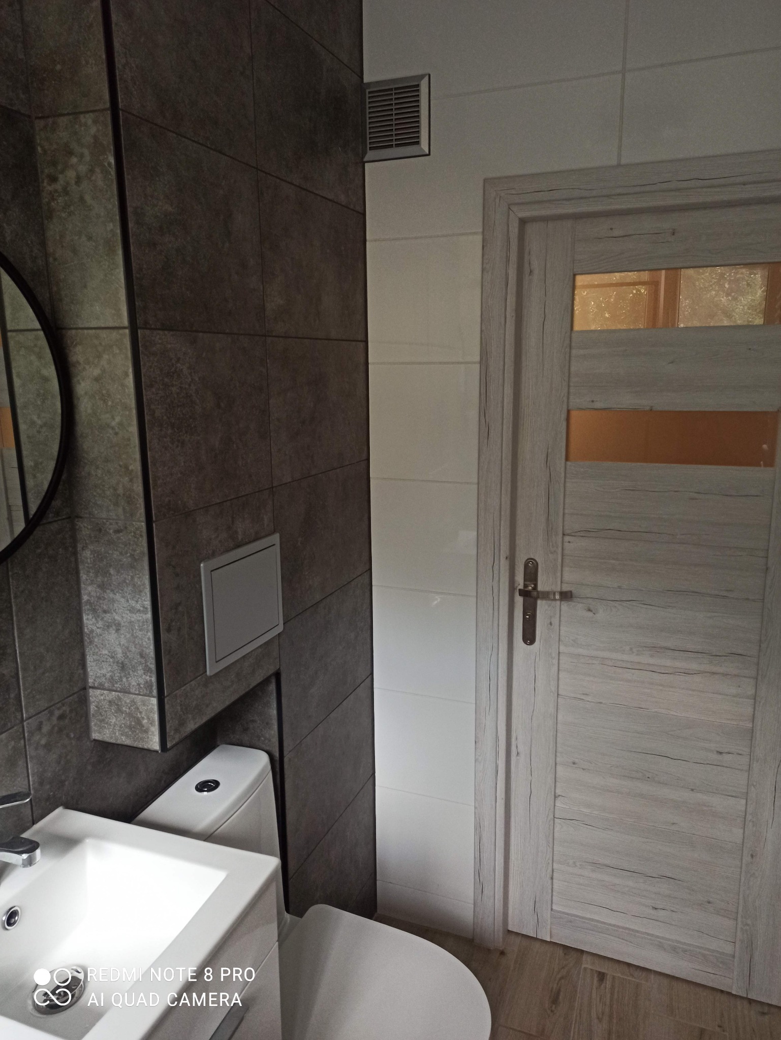 Fragment wykończonej łazienki z ciemnoszarą płytką imitującą kamień, białą ceramiką sanitarną i drewnopodobnymi drzwiami z szybą, widoczna kratka wentylacyjna na ścianie.