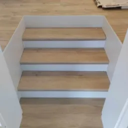 Wykończone schody z płytek imitujących drewno, białe obramowanie stopni, widoczne fragmenty podłogi i przygotowania do dalszych prac remontowych.