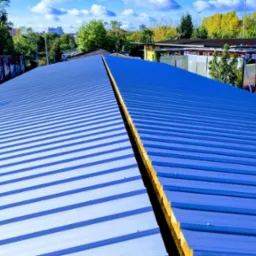 Niebieski dach z blachy trapezowej z widocznym łączeniem kalenicowym, w tle zabudowania i drzewa pod błękitnym niebem.