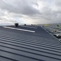 Montaż dachu z blachy trapezowej na konstrukcji stalowej, widoczne kominki wentylacyjne, pochmurne niebo.