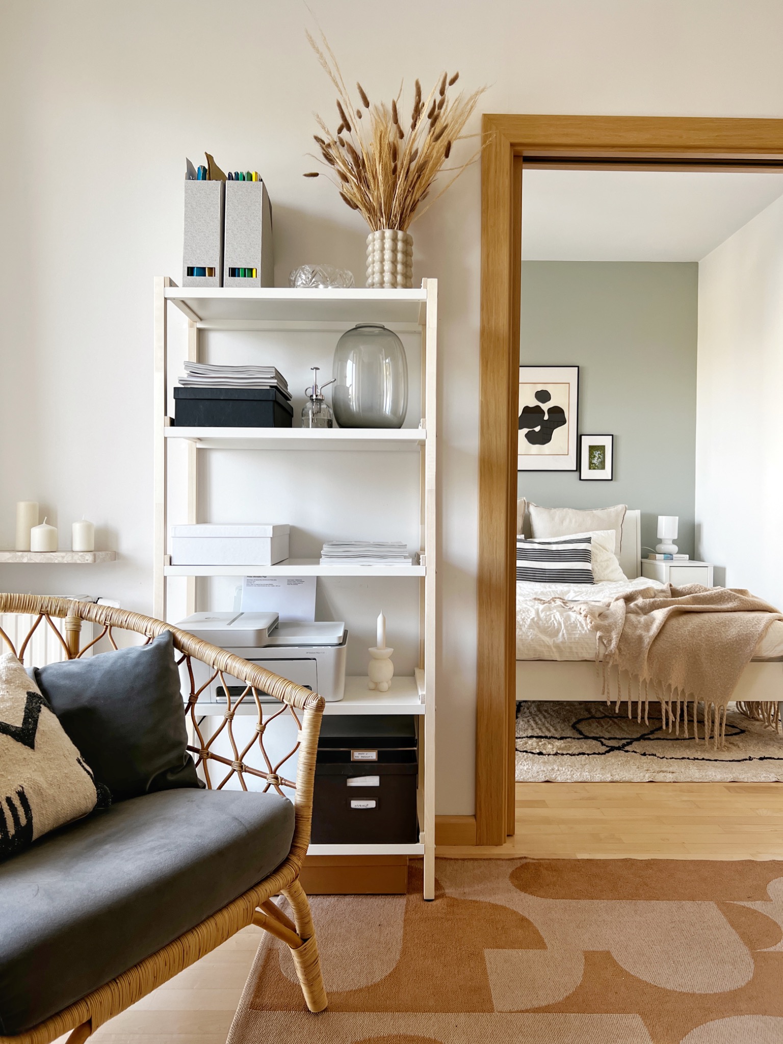 Wnętrze mieszkania z widokiem na sypialnię przez drewniane drzwi. Na pierwszym planie regał z dekoracjami i wiklinowy fotel z poduszkami. Na podłodze wzorzysty dywan w odcieniach brązu.