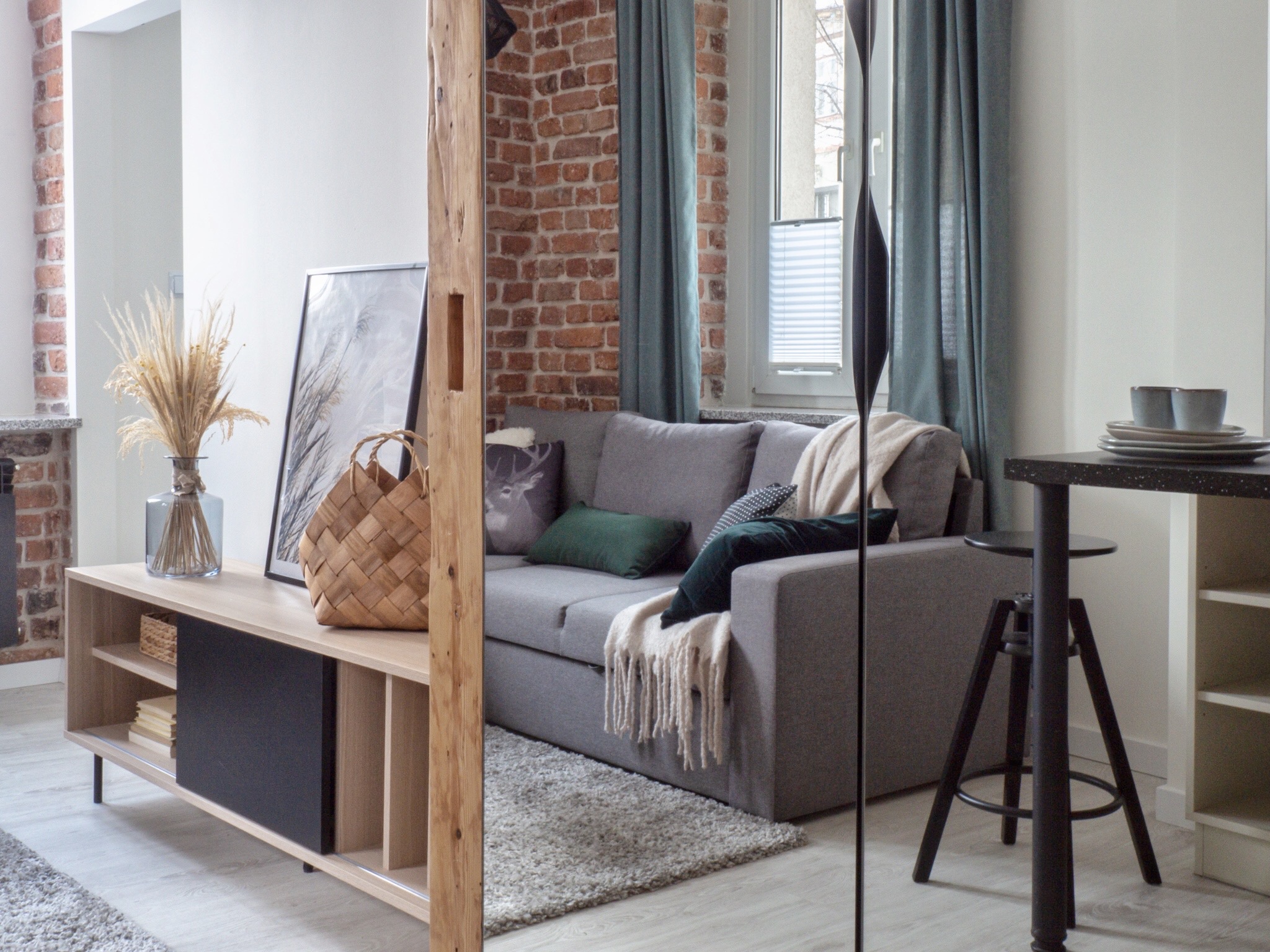 Jasnoszara sofa z poduszkami i pledem w salonie z ceglaną ścianą, drewnianym regałem z dekoracjami i stołem barowym z krzesłem, widok na okno z żaluzjami.