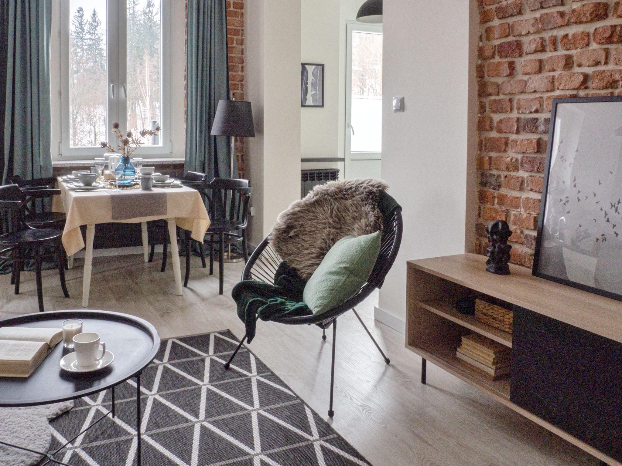 Jasne wnętrze z ceglaną ścianą, okrągłym fotelem z futrzanym pledem i zieloną poduszką, stolikiem kawowym z książką i filiżanką oraz stołem jadalnym nakrytym do posiłku.