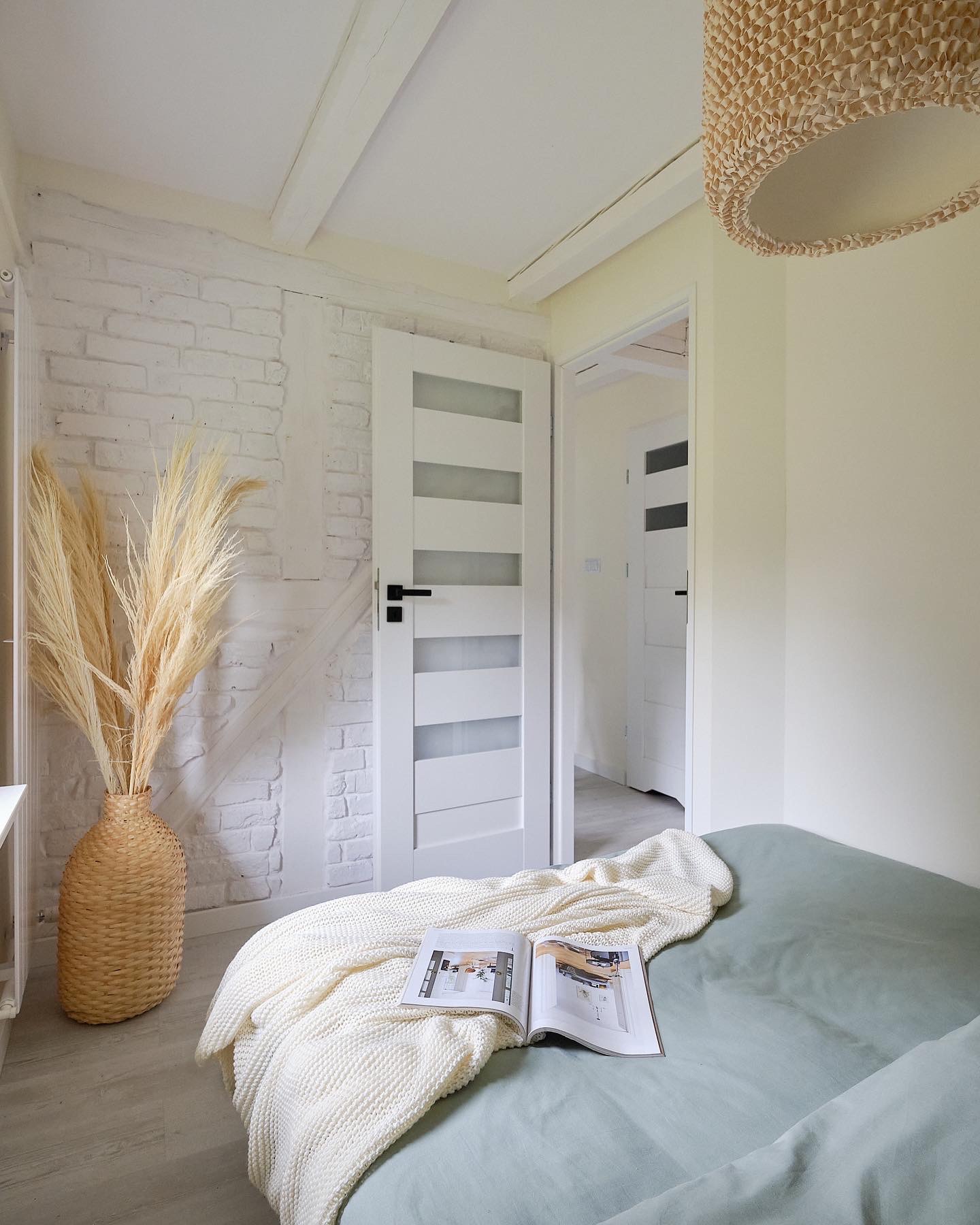 Jasne wnętrze sypialni w Warszawie z dekoracyjnym wazonem z trawą pampasową na tle białej ceglanej ściany, białe drzwi z przeszkleniami i otwarty magazyn na łóżku z zieloną pościelą.