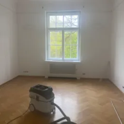 Pokój po pracach, widoczny parkiet ułożony w jodełkę, odkurzacz przemysłowy Festool, białe ściany z dekoracyjnym gzymsem pod sufitem, okno z widokiem na drzewa.