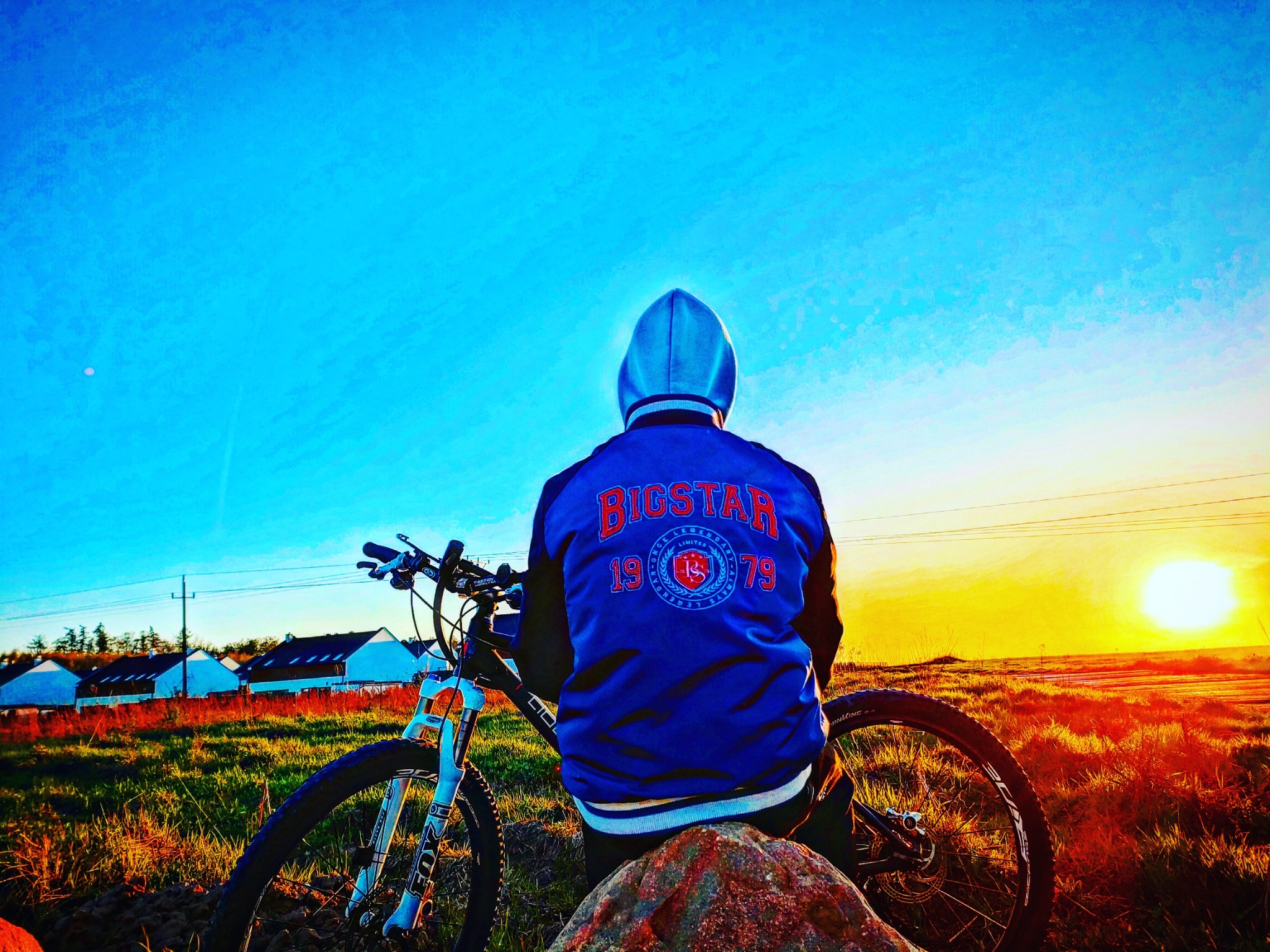 Mężczyzna w niebieskiej bluzie z kapturem siedzi na kamieniu obok roweru górskiego na tle zachodzącego słońca i rzędu białych domów.