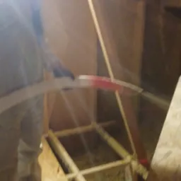 Pracownik w kombinezonie ochronnym i masce tnie nożem izolację w trakcie prac na poddaszu, widoczna drewniana konstrukcja dachu i fragment oświetlenia.