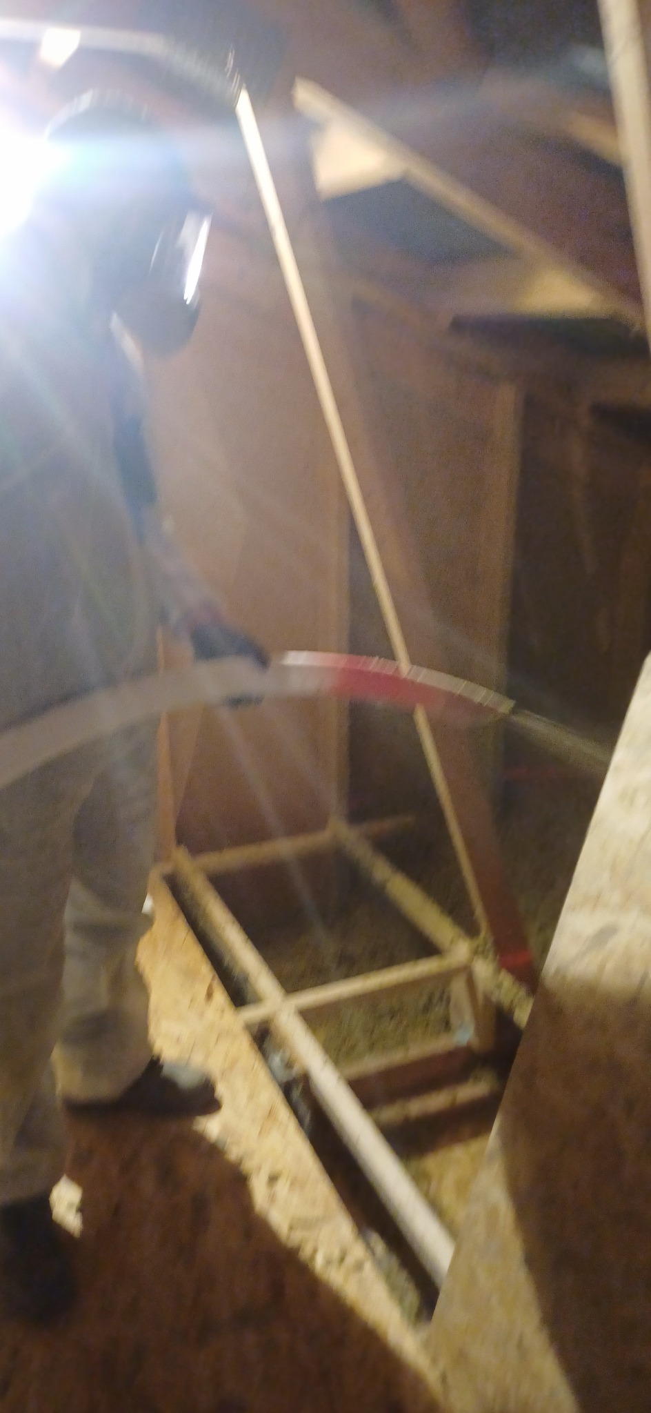 Pracownik w kombinezonie ochronnym i masce tnie nożem izolację w trakcie prac na poddaszu, widoczna drewniana konstrukcja dachu i fragment oświetlenia.