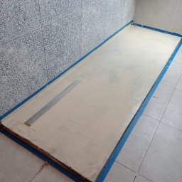 Łukasz Bartuzel - Przygotowany brodzik z odpływem liniowym, zabezpieczony niebieską taśmą malarską, w trakcie wylewania posadzki w łazience z dekoracyjnymi płytkami ściennymi.
