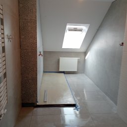 Łukasz Bartuzel - Wnętrze łazienki w trakcie prac wykończeniowych, widoczne świeżo wylane podłoże pod posadzkę, zabezpieczone taśmą malarską, płytki na ścianach i podłodze, oraz grzejnik na ścianie i okno dachowe.