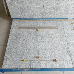 Łukasz Bartuzel - Układanie płytek podłogowych z geometrycznym wzorem, z użyciem poziomicy i klipsów poziomujących, zabezpieczone niebieską taśmą malarską.