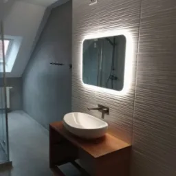 Nowoczesna łazienka na poddaszu z umywalką nablatową na drewnianej szafce, podświetlanym lustrem i płytkami ściennymi o falistej strukturze.