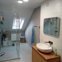 Łukasz Bartuzel - Jasna łazienka na poddaszu z kabiną prysznicową typu walk-in, umywalką nablatową na drewnianej szafce i lustrem z zaokrąglonymi rogami na ścianie z dekoracyjnymi płytkami.