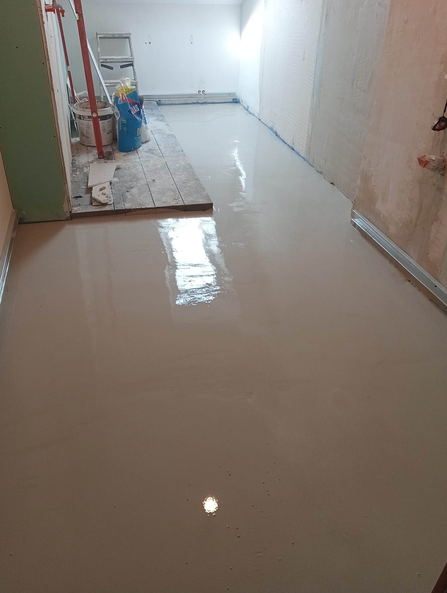 Wylewka samopoziomująca w trakcie schnięcia, widoczne narzędzia i materiały budowlane w tle, przygotowane podłoże z fragmentami desek.