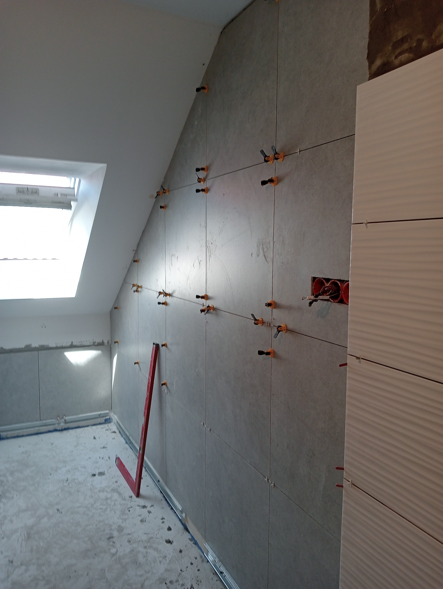 Montaż szarych płytek ściennych w łazience na poddaszu, widoczne poziomice, krzyżyki dystansowe i puszka elektryczna.