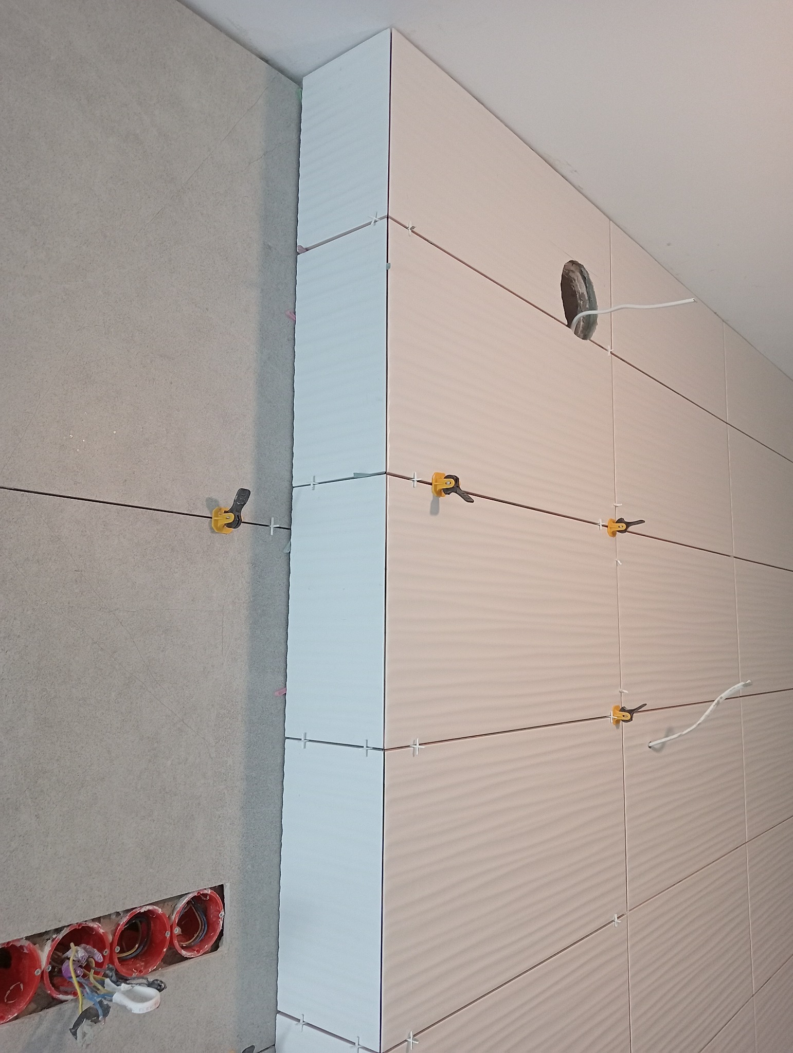 Ściana w trakcie wykańczania płytkami ceramicznymi o falistej strukturze, z widocznymi krzyżykami dystansowymi i systemem poziomowania płytek, obok ściana z widocznymi puszkami elektrycznymi.