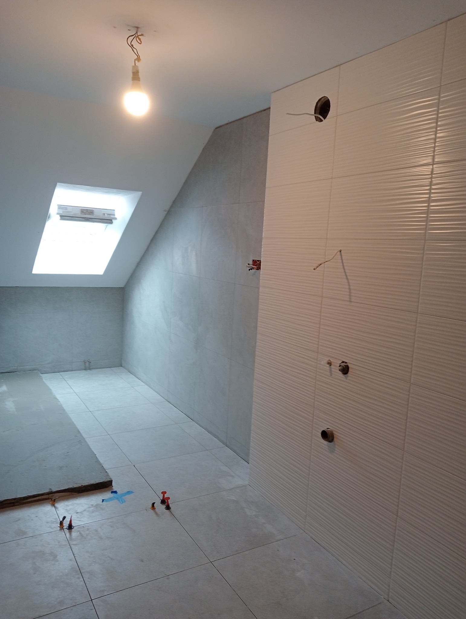 Poddasze łazienki w trakcie prac wykończeniowych: ułożone płytki podłogowe z systemem poziomowania, częściowo położone płytki ścienne z fakturą, widoczne otwory na instalacje wodne i elektryczne...