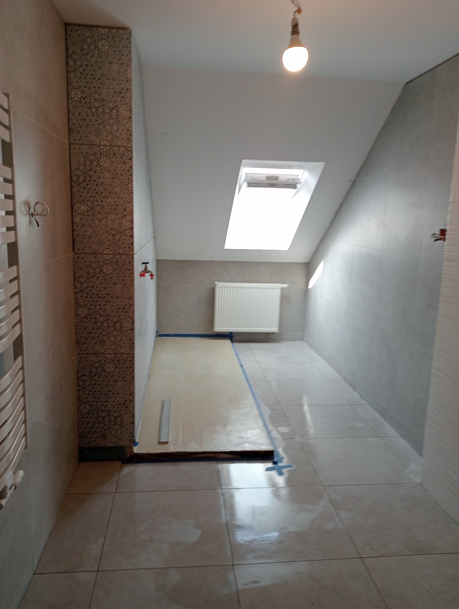 Wnętrze łazienki w trakcie prac wykończeniowych, widoczne świeżo wylane podłoże pod posadzkę, zabezpieczone taśmą malarską, płytki na ścianach i podłodze, oraz grzejnik na ścianie i okno dachowe.