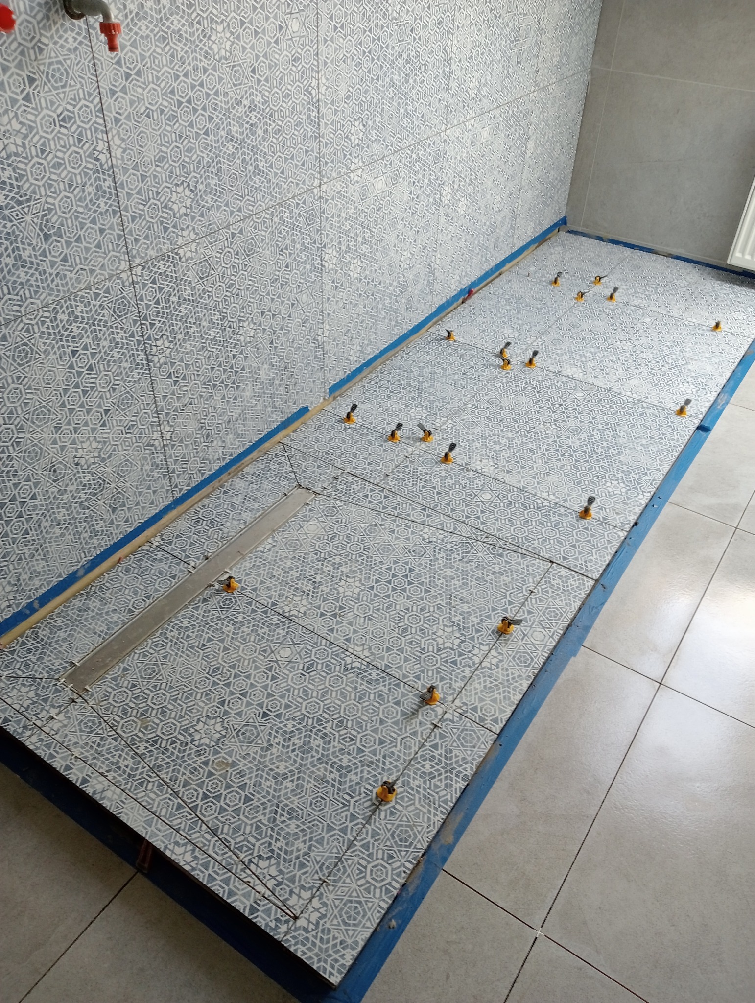 Podczas układania płytek z geometrycznym wzorem w łazience, widoczny odpływ liniowy i system poziomowania płytek z pomarańczowymi klinami.