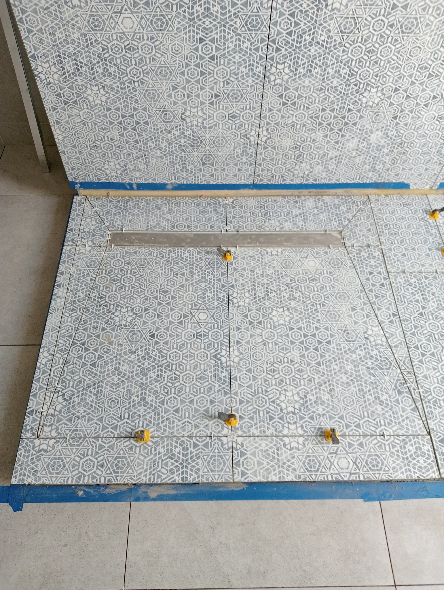 Układanie płytek podłogowych z geometrycznym wzorem, z użyciem poziomicy i klipsów poziomujących, zabezpieczone niebieską taśmą malarską.