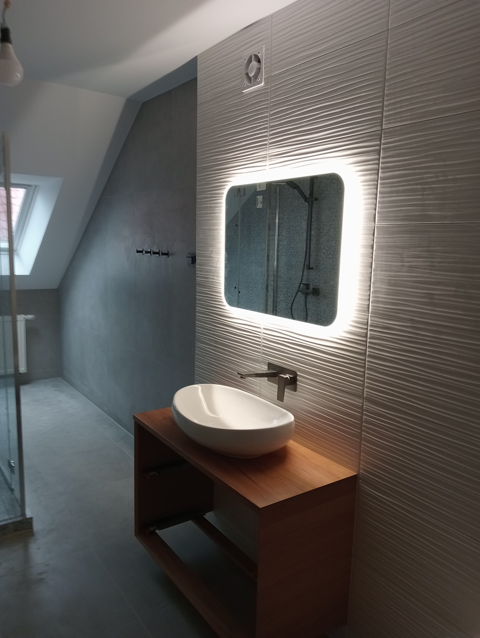 Nowoczesna łazienka na poddaszu z umywalką nablatową na drewnianej szafce, podświetlanym lustrem i płytkami ściennymi o falistej strukturze.