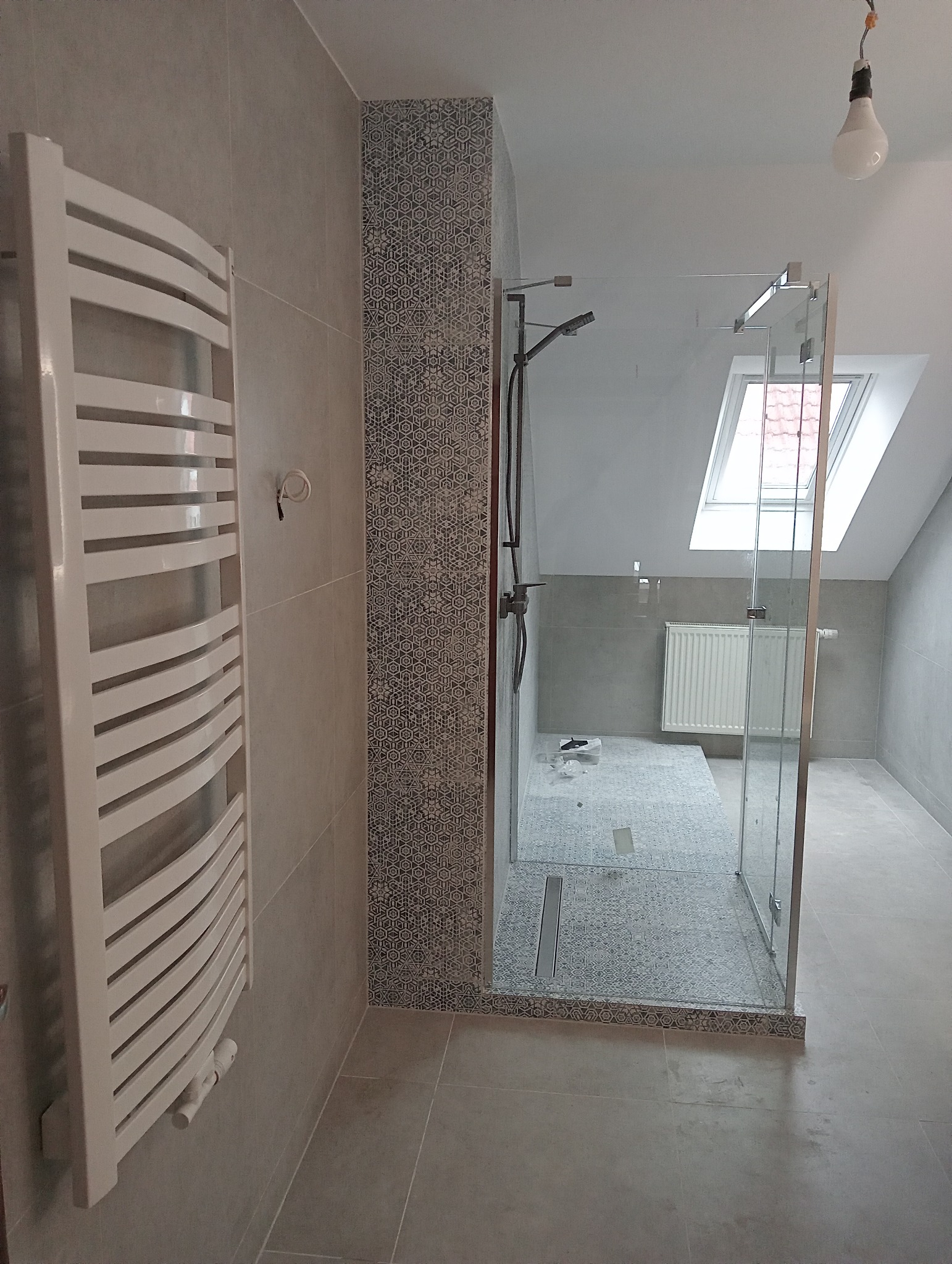 Szara łazienka w trakcie remontu z kabiną prysznicową typu walk-in, grzejnikiem drabinkowym i dekoracyjną mozaiką na obudowie prysznica, widoczne narzędzia i niedokończone prace.