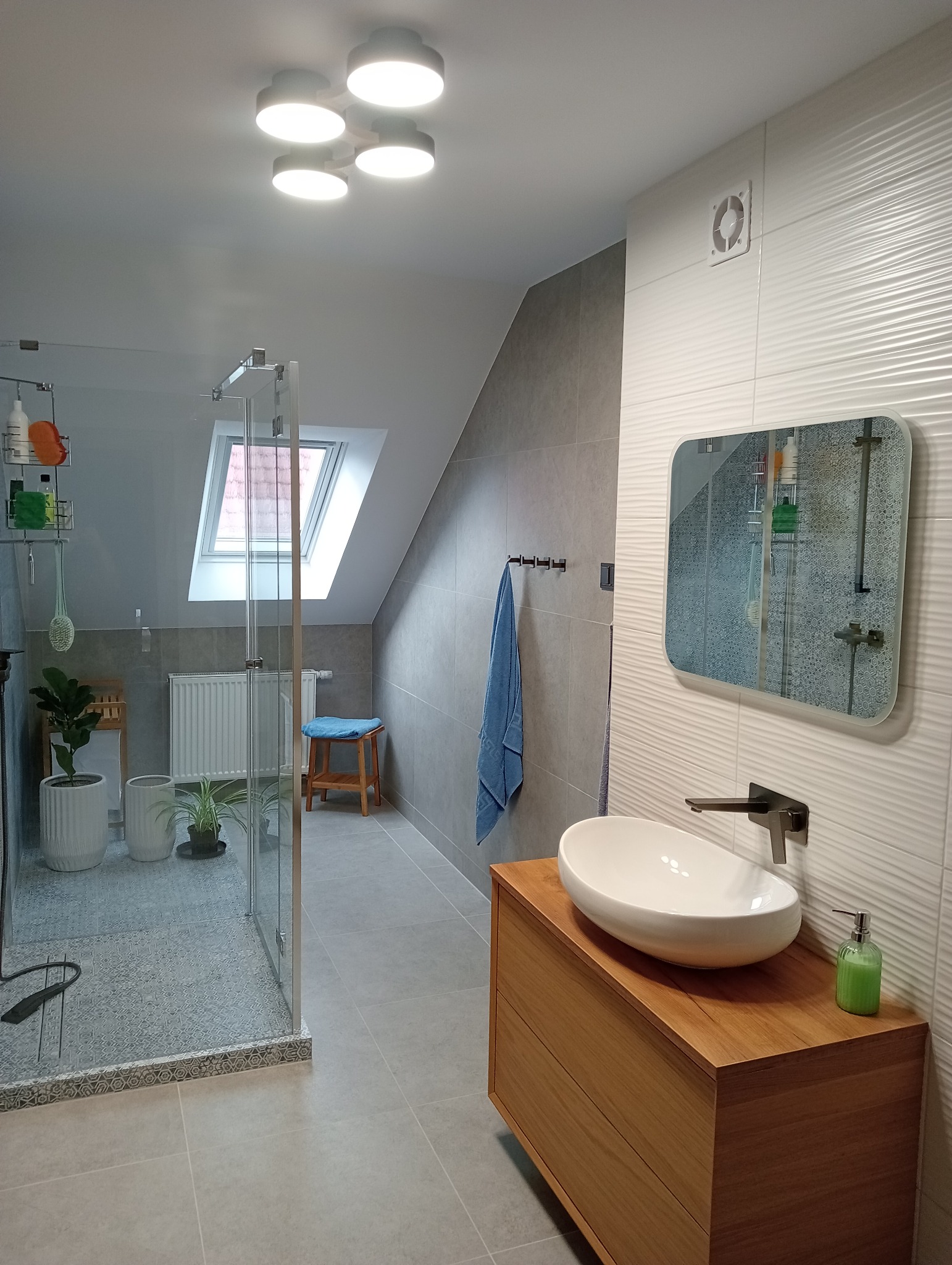 Jasna łazienka na poddaszu z kabiną prysznicową typu walk-in, umywalką nablatową na drewnianej szafce i lustrem z zaokrąglonymi rogami na ścianie z dekoracyjnymi płytkami.