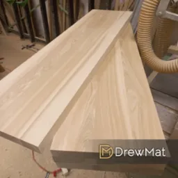 Dwa blaty z jasnego drewna leżące jeden na drugim w warsztacie, widoczne słoje i faktura drewna, w tle rura od odciągu trocin i elementy konstrukcyjne, w dolnym prawym rogu logo firmy.