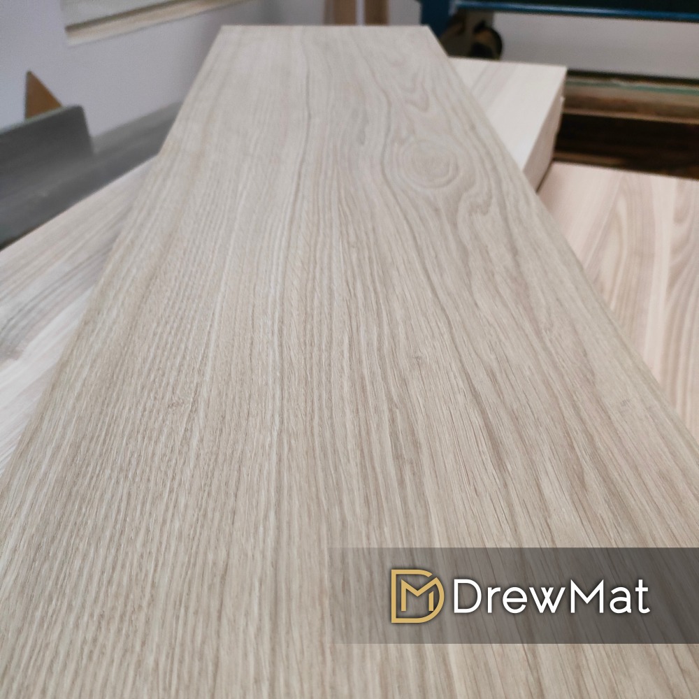 Deska z jasnego drewna dębowego z widocznym usłojeniem, ułożona na stole w warsztacie, w tle maszyny stolarskie, znak wodny z nazwą firmy.