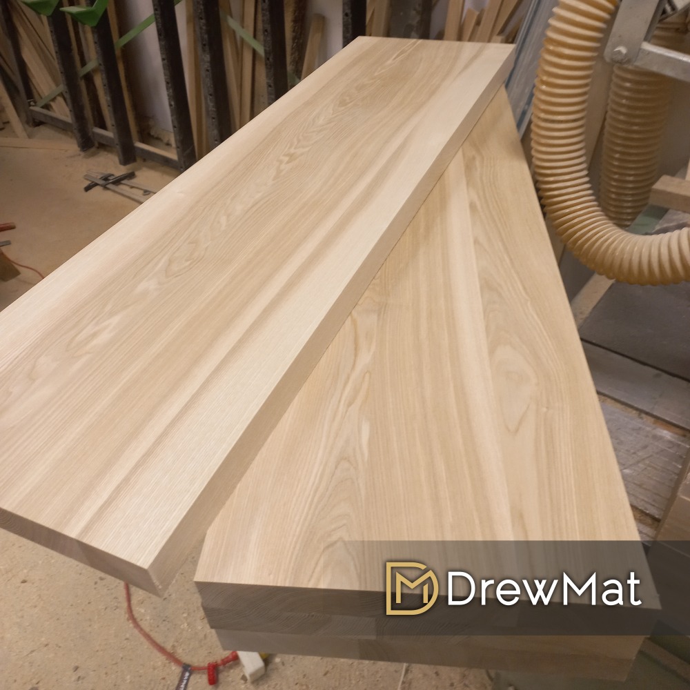 Dwa blaty z jasnego drewna leżące jeden na drugim w warsztacie, widoczne słoje i faktura drewna, w tle rura od odciągu trocin i elementy konstrukcyjne, w dolnym prawym rogu logo firmy.