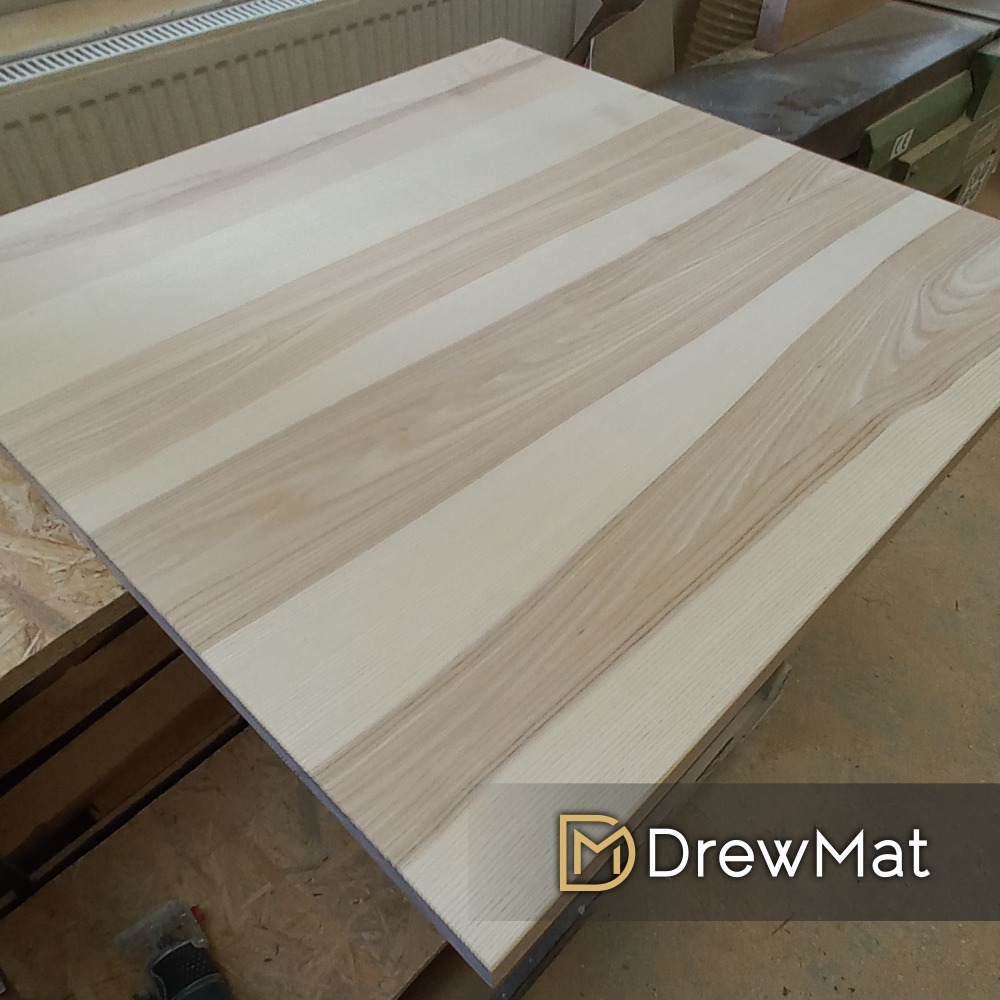 Kwadratowy blat z jasnego drewna klejonego warstwowo, z widocznym usłojeniem i delikatnymi różnicami w odcieniach, umieszczony na warsztacie.